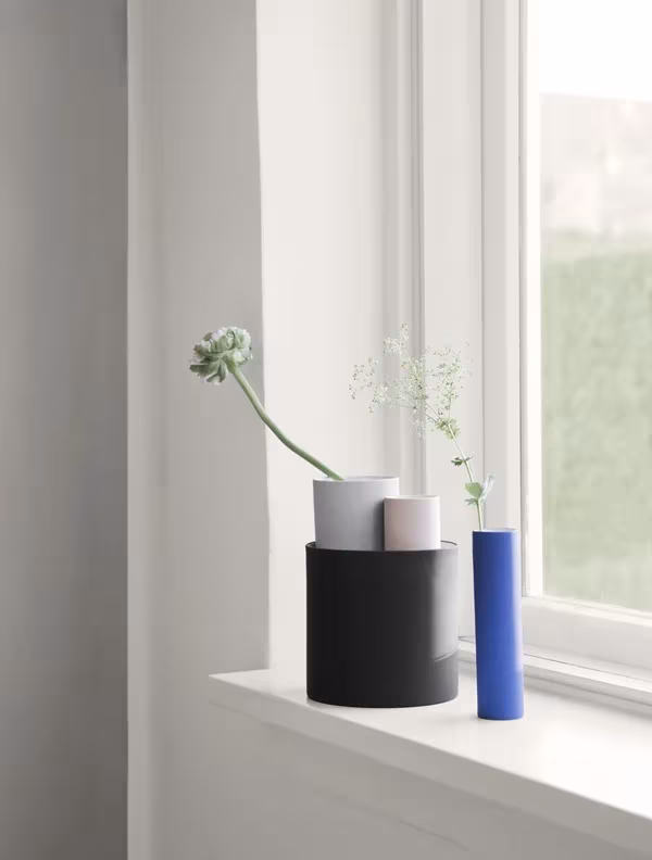 Jarrones cilíndricos apilables de Ferm Living en negro, blanco y azul con flores, en el alféizar de una ventana