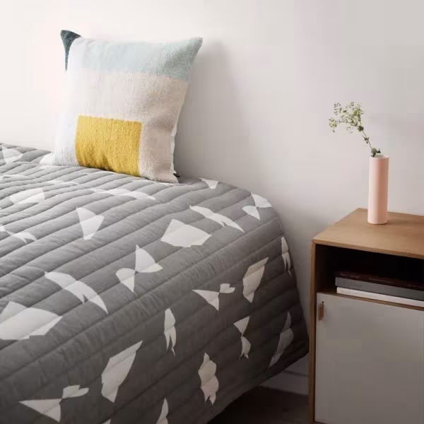 Dormitorio con colcha gris de motivos geométricos, cojín de punto de Ferm Living y mesilla de madera con jarrón rosa