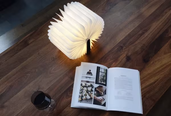 Lámpara Lumio encendida sobre una mesa de madera oscura junto a un libro abierto y una copa de vino