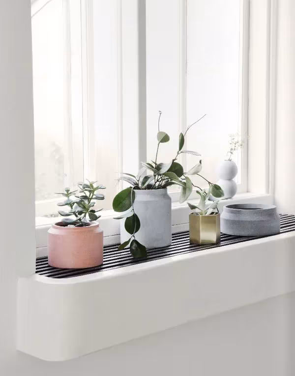 Macetas de cerámica de Ferm Living en rosa, gris y dorado con plantas sobre un alféizar blanco junto a la ventana
