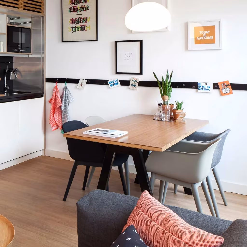 Zona de comedor de un Loft ZOKU en Ámsterdam con mesa de madera, sillas grises, cocina blanca y láminas en la pared