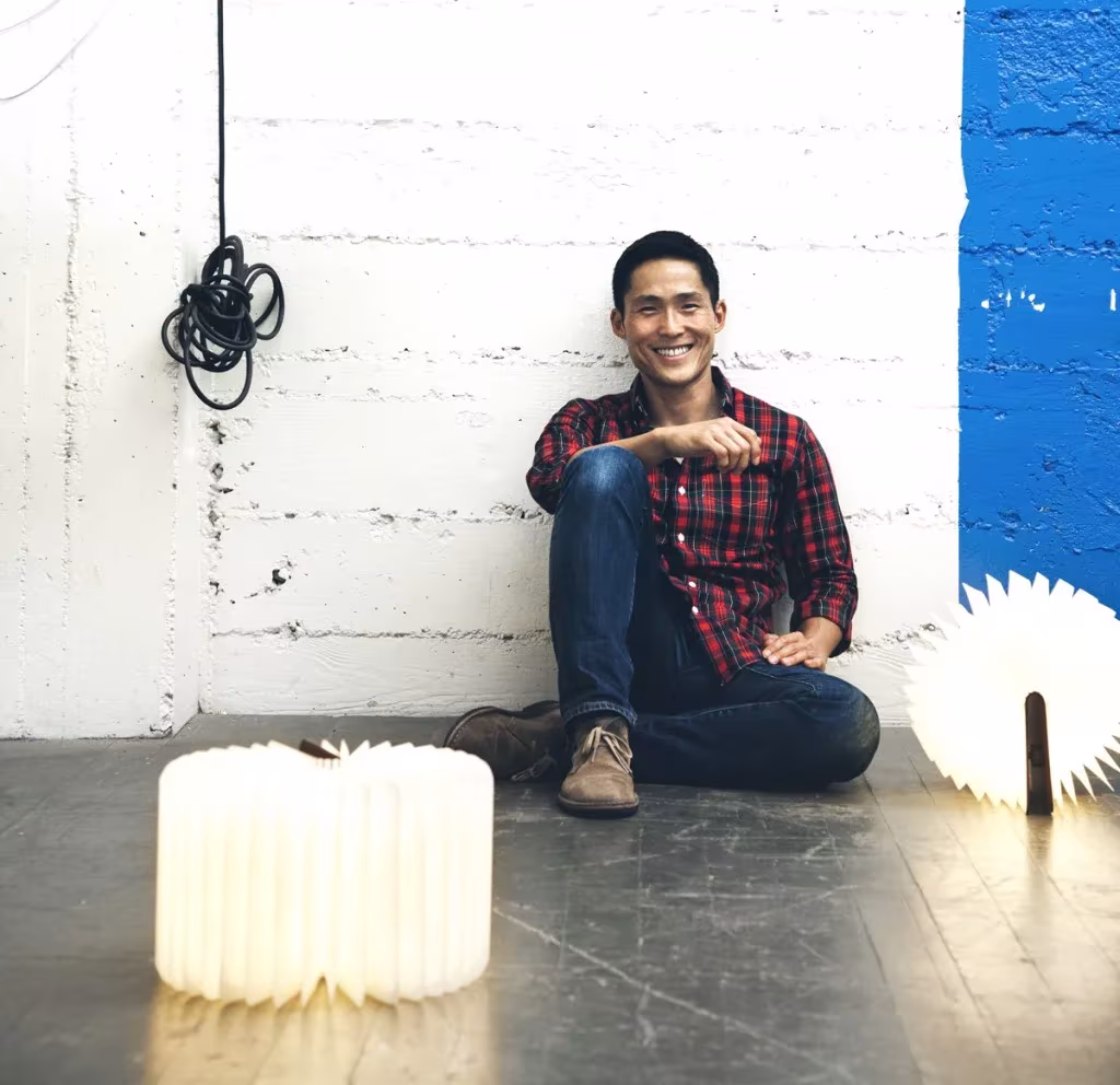 Hombre sonriente sentado en el suelo junto a una pared encalada con dos lámparas Lumio encendidas a sus lados