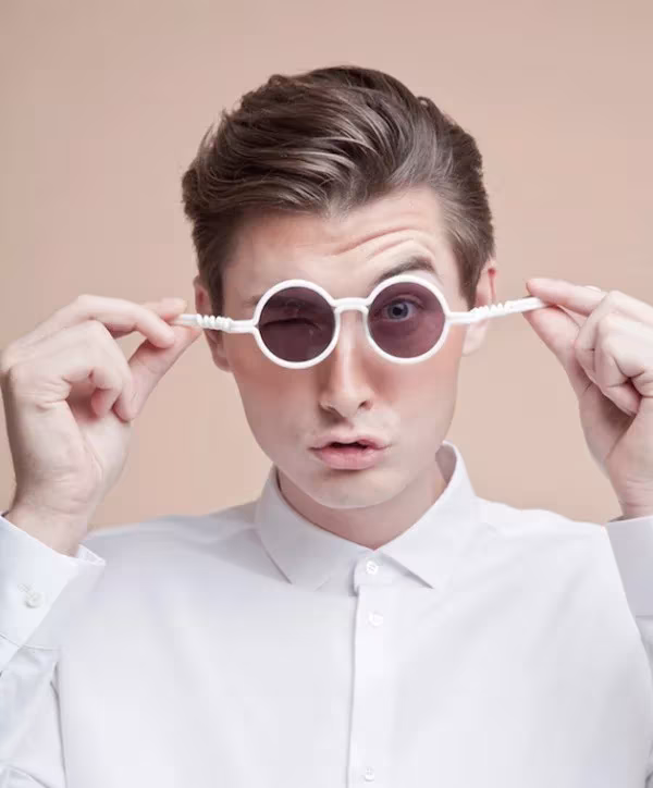 Hombre con camisa blanca ajustándose unas gafas MONO redondas blancas de lentes rosadas sobre fondo rosa