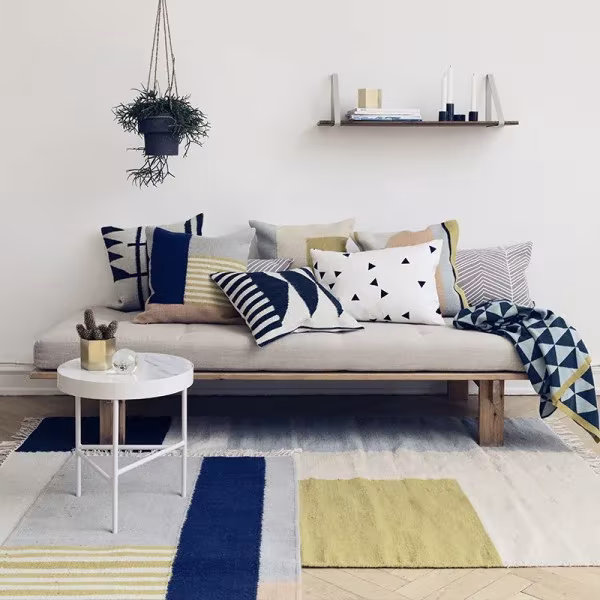 Salón con sofá de madera, cojines geométricos y manta de triángulos de Ferm Living, alfombra a bloques de color y maceta colgante