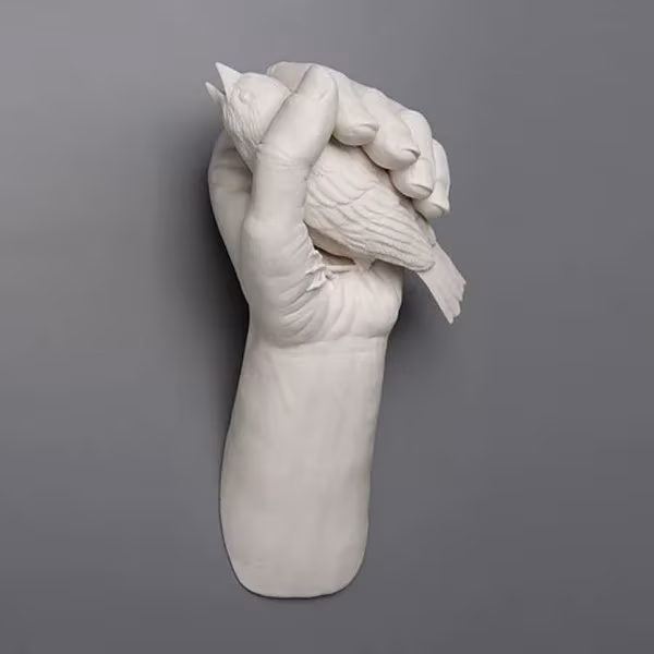 Escultura de porcelana blanca de Kate MacDowell: mano y antebrazo que aprietan un pájaro, sobre fondo gris