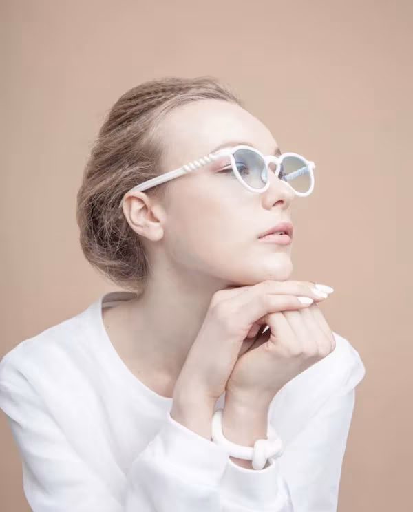 Mujer con jersey blanco y gafas MONO blancas apoyando la barbilla en las manos sobre fondo rosado