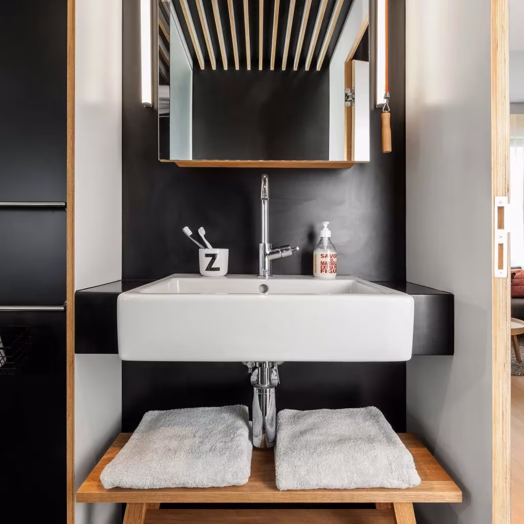 Lavabo blanco sobre encimera negra y taburete de madera con toallas en el baño de un Loft ZOKU de Ámsterdam