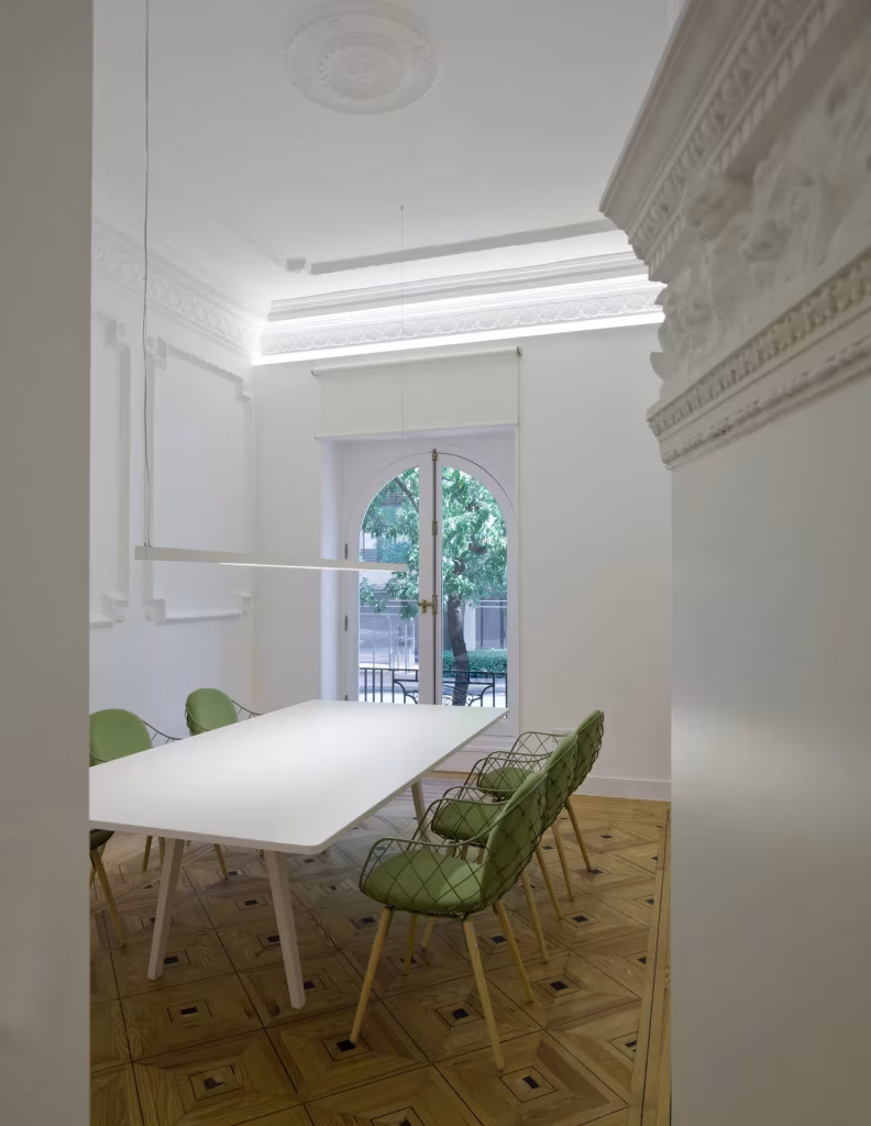 Sala de reuniones blanca con mesa larga y sillas verdes junto a un ventanal en arco, oficina de ÁBATON
