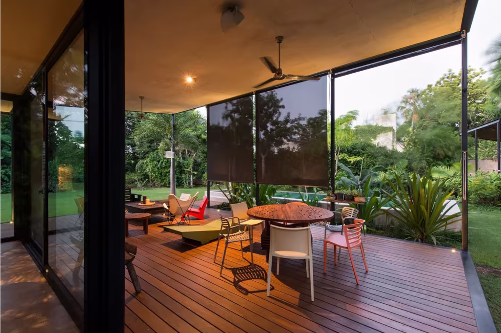 Terraza cubierta de la Casa Itzimná con tarima de madera, mesa redonda y sillas de colores abierta al jardín