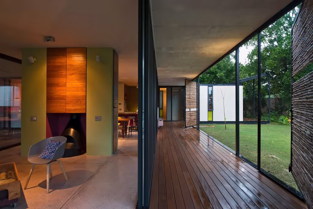 Pasillo de madera de la Casa Itzimná junto a un ventanal al césped, con chimenea revestida de madera y butaca