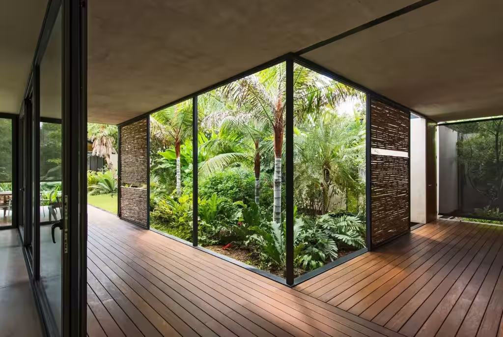 Esquina de vidrio sin montante de la Casa Itzimná con tarima de madera y un patio de palmeras y helechos