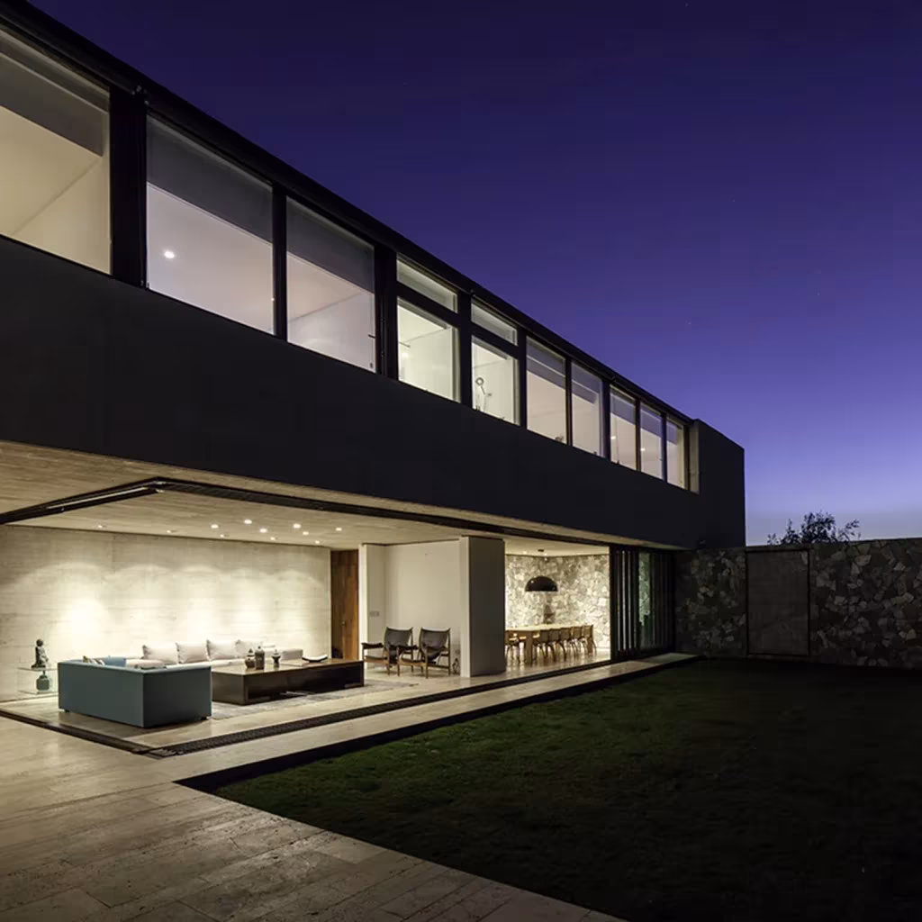 Casa SH de 01ARQ al anochecer, volumen superior oscuro en voladizo sobre un salón iluminado abierto al césped