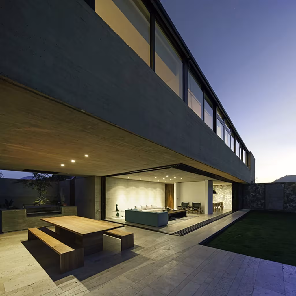 Terraza de la Casa SH con mesa y bancos de madera bajo el voladizo de hormigón, y salón iluminado detrás