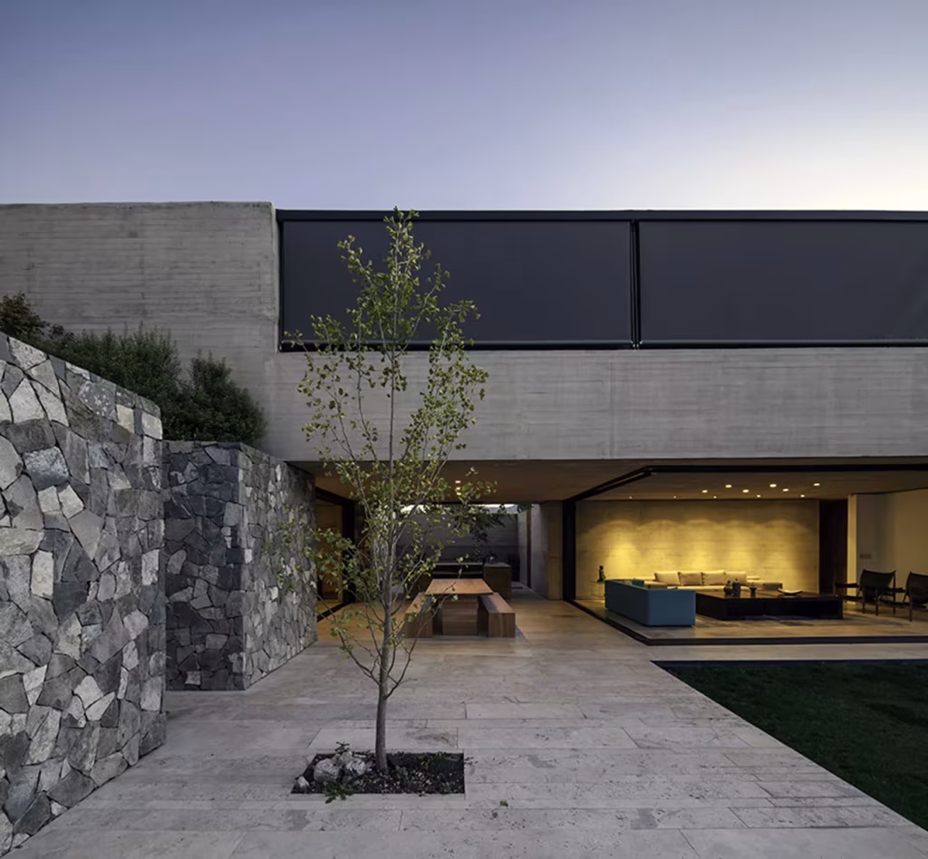 Patio de acceso de la Casa SH con muro de mampostería de piedra, árbol joven y salón acristalado al fondo
