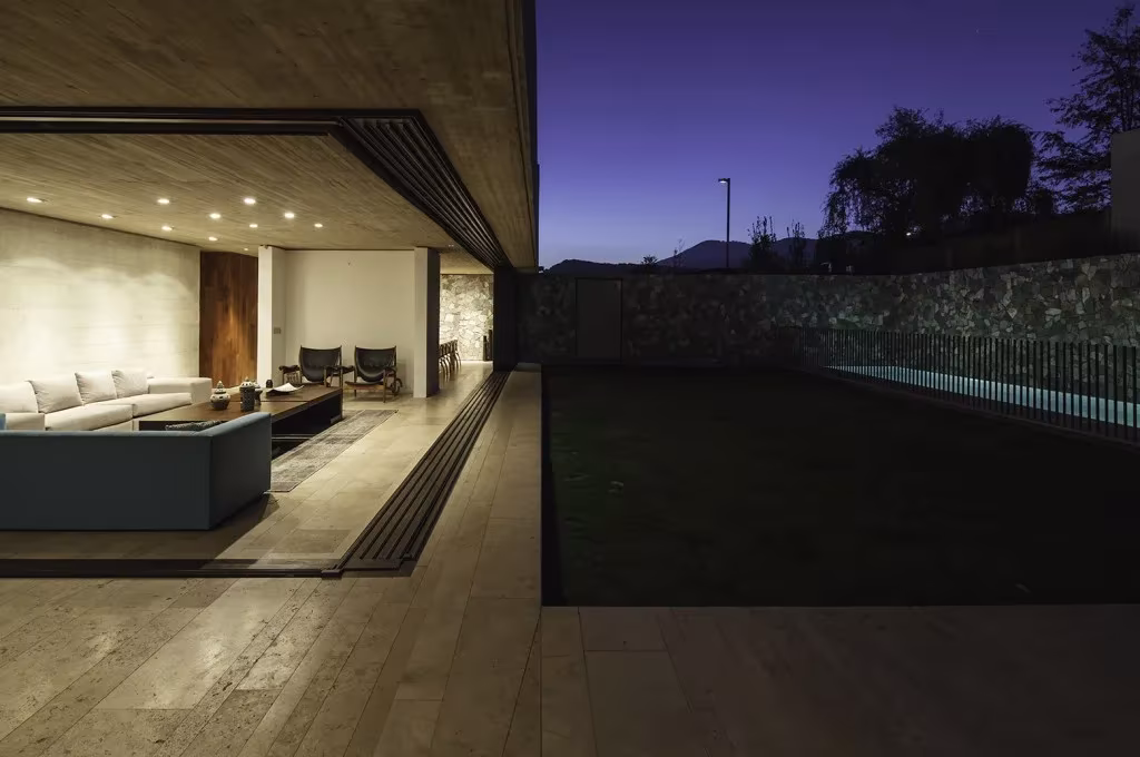 Salón de la Casa SH abierto de noche al jardín, con sofá blanco, suelo de piedra y muro de mampostería