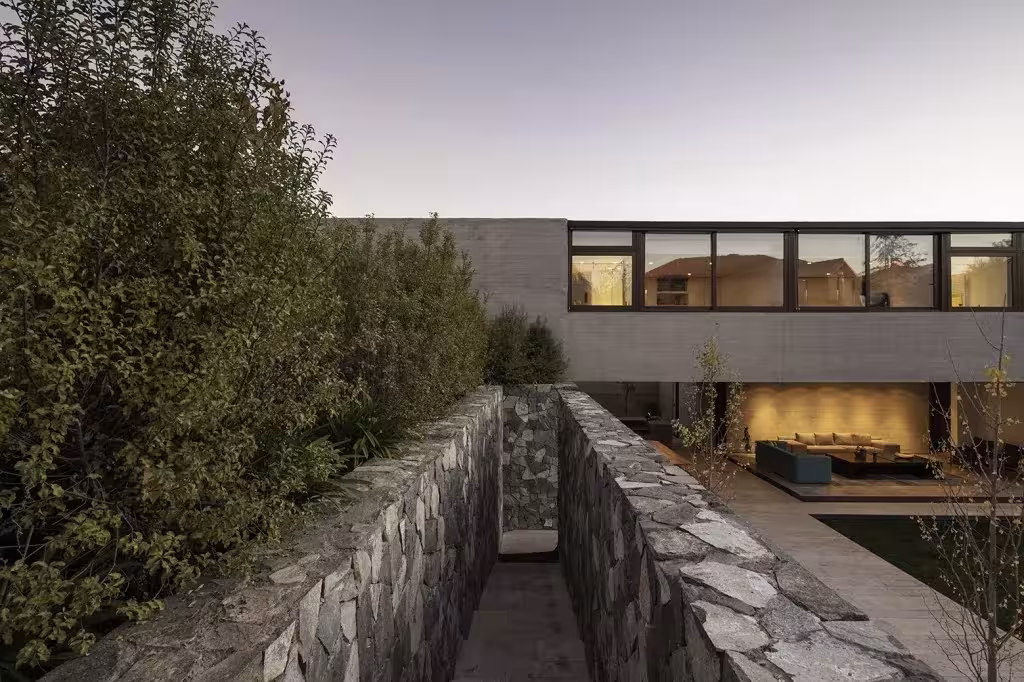 Pasaje entre muros de piedra hacia la Casa SH, con vegetación densa y ventanas iluminadas en la planta alta