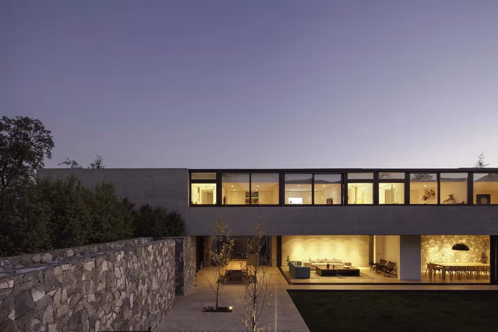 Casa SH al crepúsculo con fachada de hormigón, ventanas corridas iluminadas y patio inferior junto a un muro de piedra