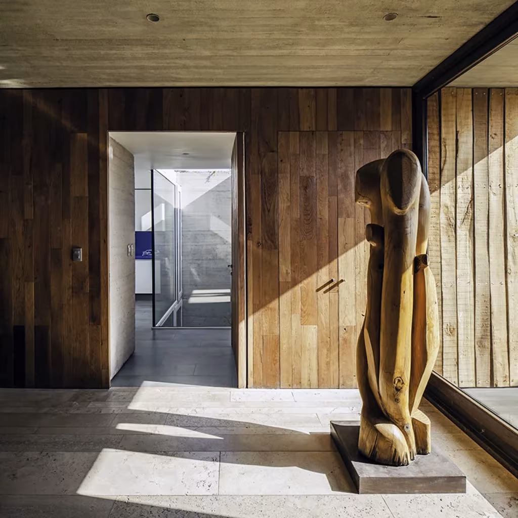 Vestíbulo de la Casa SH con pared de tablones de madera, puerta acristalada y escultura de madera tallada