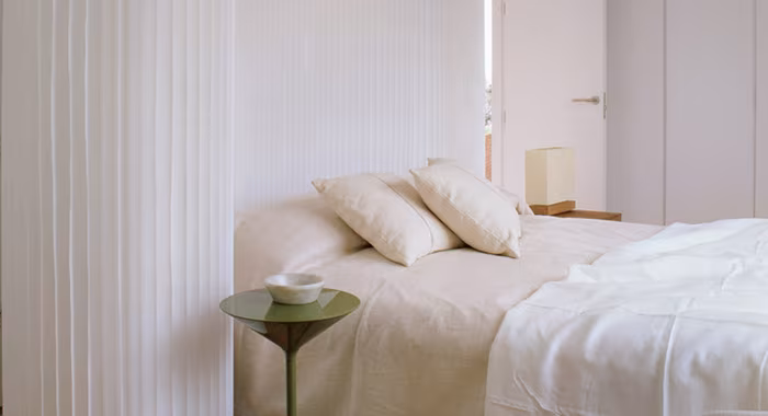 Dormitorio con cama de ropa blanca y un biombo Molo de pliegues claros como separador, proyecto de Batavia