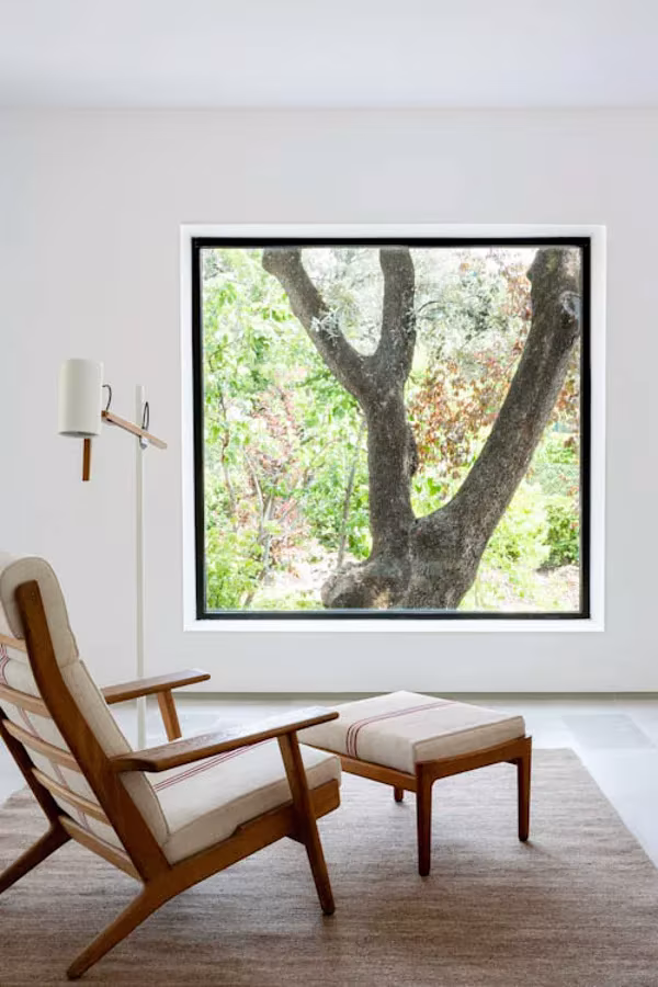 Butaca de madera con cojines blancos y reposapiés junto a una gran ventana cuadrada que enmarca un árbol, en La Moraleja