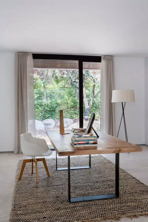 Despacho con mesa de madera maciza, silla blanca, lámpara de pie y alfombra de yute frente a un ventanal al jardín