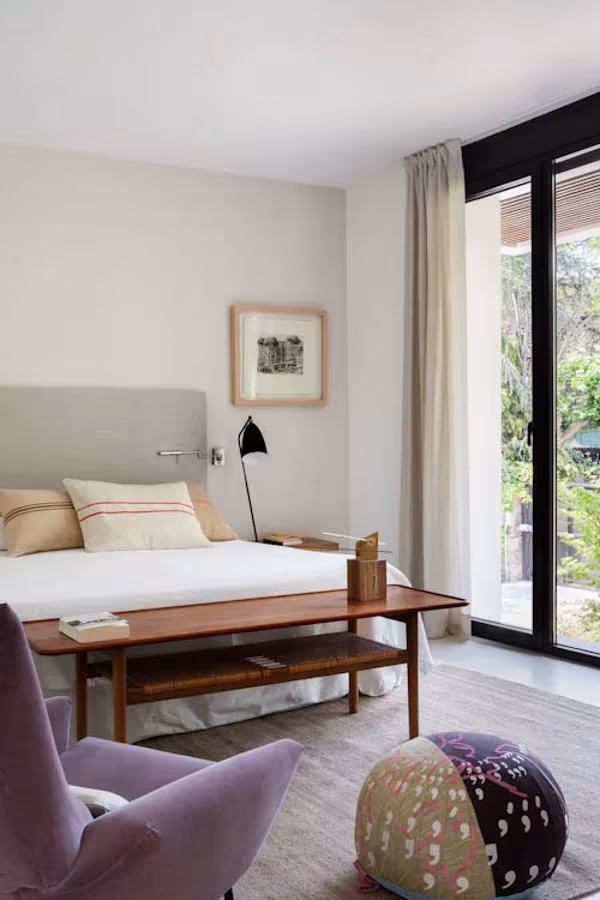 Dormitorio luminoso de BATAVIA con cama blanca, banco de madera, butaca malva y puf estampado junto al ventanal