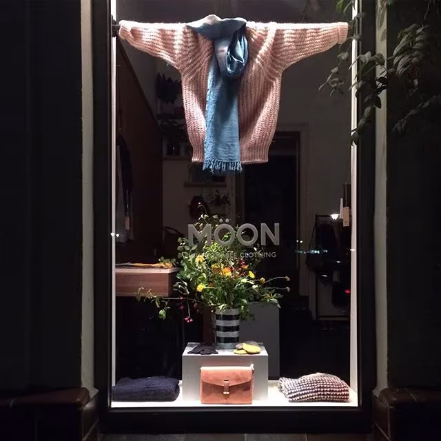 Escaparate nocturno de la tienda Möon en Berlín con un jersey rosa colgado, flores silvestres y bolsos de piel