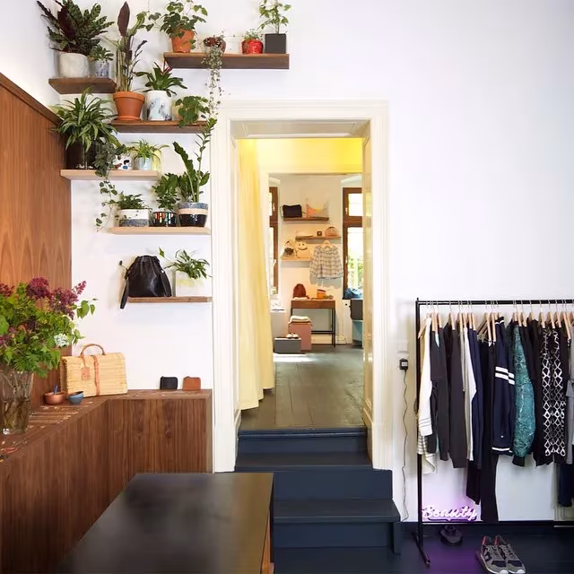 Interior de Möon con estantes de plantas en pared blanca, mueble de nogal, bolsos y un perchero de ropa