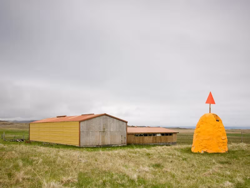 Naves agrícolas de chapa amarilla y gris en una pradera islandesa junto a un montón de heno con baliza naranja