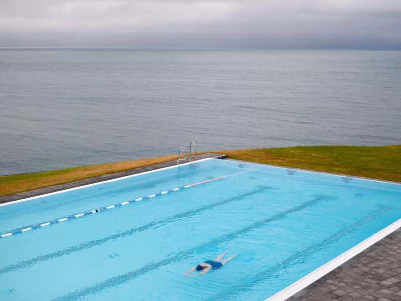 Bañista nadando en una piscina exterior azul al borde del mar bajo un cielo gris en Islandia