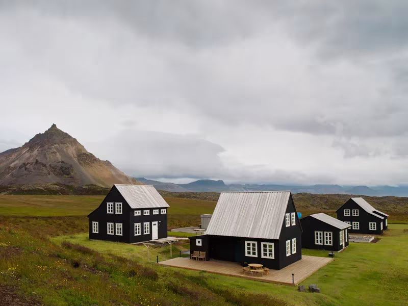 Casas negras de tejado gris en un prado verde al pie de una montaña en Islandia, serie Landnemar