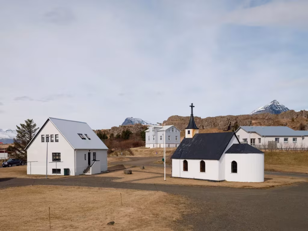 Pequeña iglesia negra con campanario junto a casas blancas en un pueblo islandés con montañas al fondo