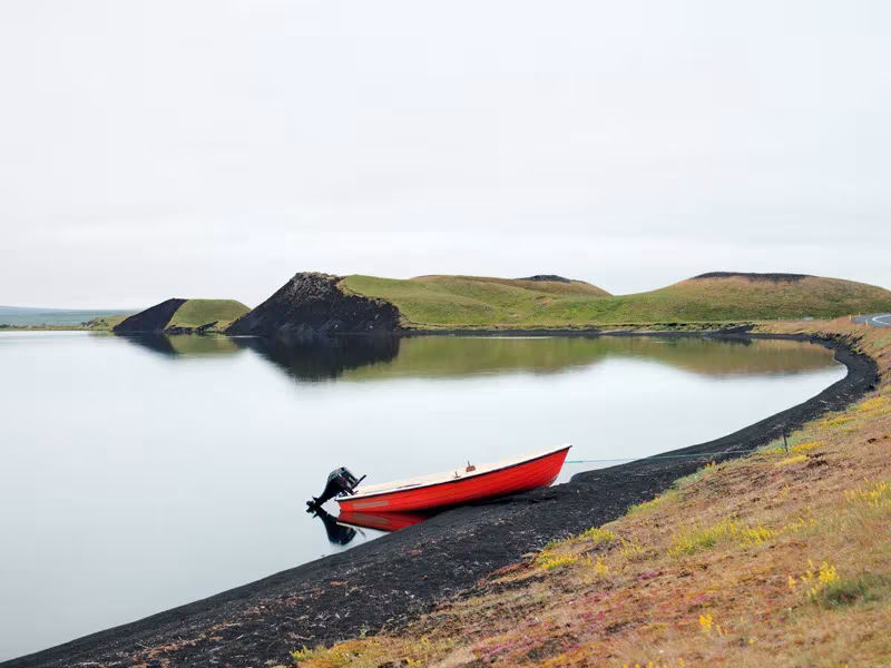 Barca roja varada en la orilla de arena negra de un lago en calma con colinas verdes en Islandia