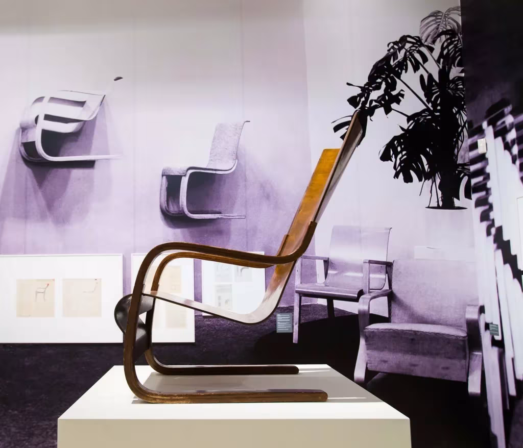Butaca de madera curvada de Alvar Aalto expuesta en CaixaForum Madrid ante una pared malva con sillas colgadas