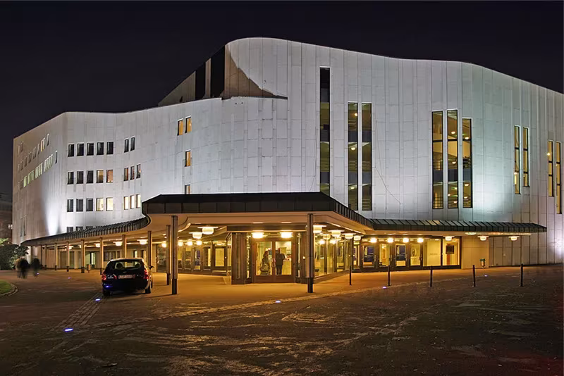 Teatro Aalto de Essen iluminado de noche, con fachada blanca curva y marquesina de entrada acristalada
