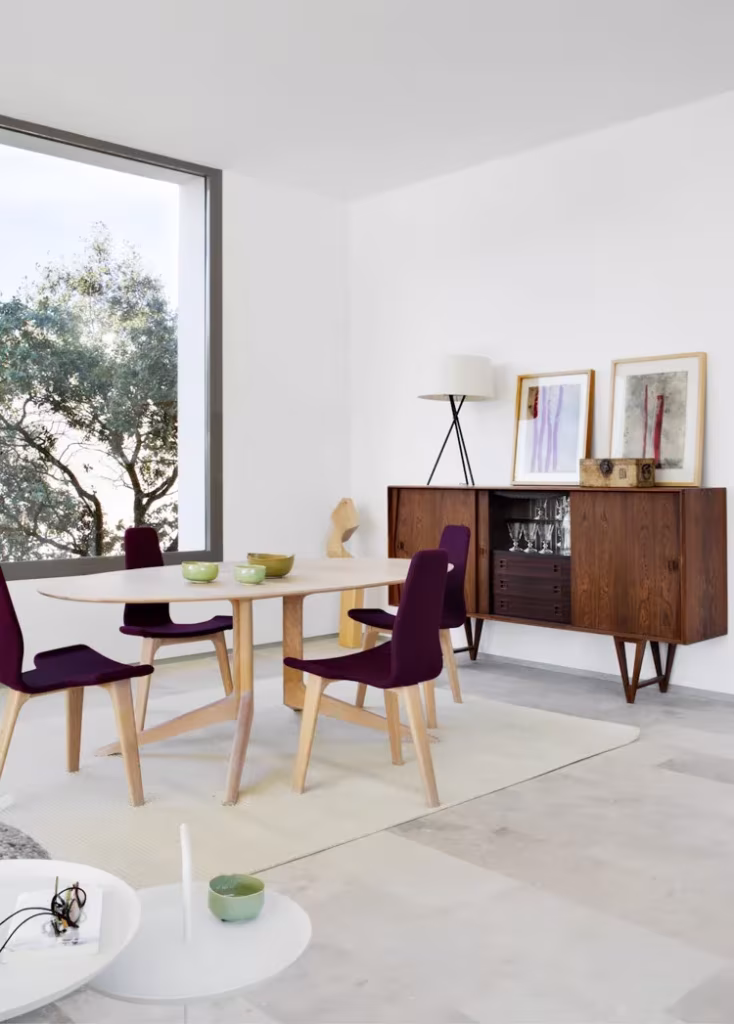 Comedor de Montealegría con mesa ovalada, sillas moradas, aparador de madera y ventanal hacia los árboles