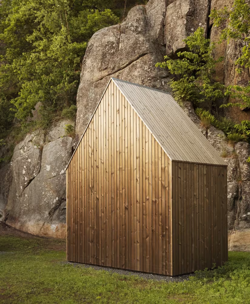 Pequeña cabaña de madera a dos aguas de Reiulf Ramstad Arkitekter junto a una gran roca con vegetación