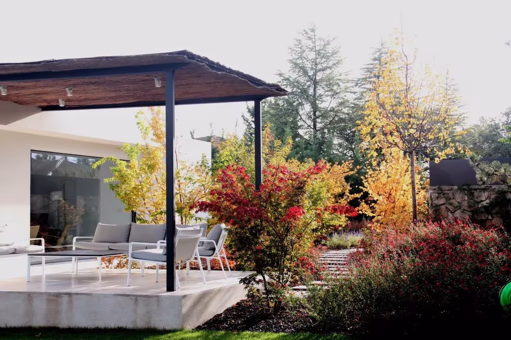 Porche con pérgola y sofás grises abierto a un jardín otoñal de arces rojos y árboles amarillos, por ÁBATON