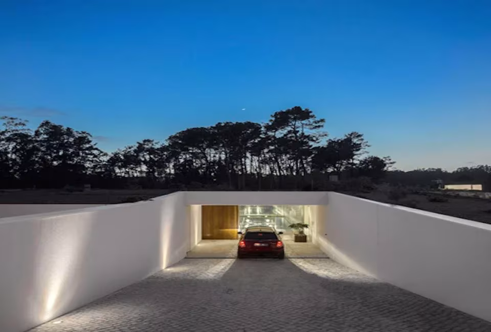 Rampa de acceso entre muros blancos hacia el garaje iluminado de la Hill Cork House al anochecer