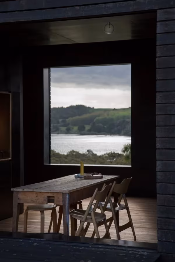 Interior oscuro de la cabaña Eyrie con mesa de madera, sillas plegables y ventanal hacia el estuario