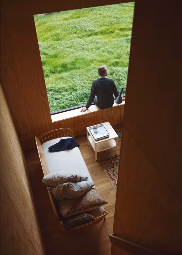 Persona sentada en el alféizar de la cabaña Eyrie mirando la hierba, con un sofá cama de madera dentro