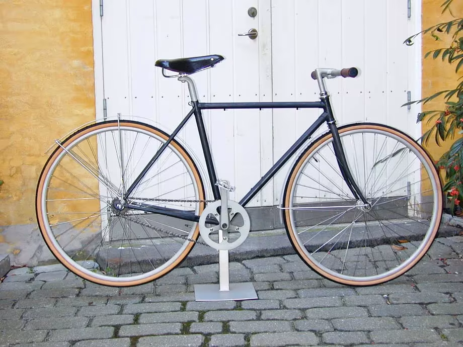 Bicicleta Sögreni Classic azul con ruedas de pared marrón ante una puerta blanca sobre adoquines