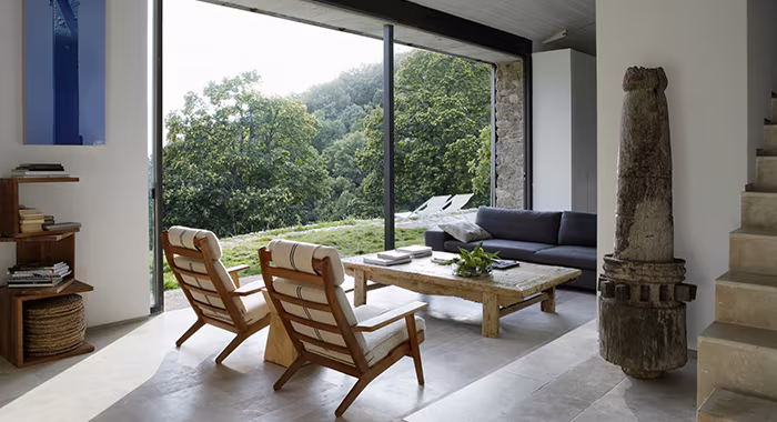 Salón con butacas de madera, sofá oscuro y gran ventanal abierto al bosque en el establo rehabilitado por ÁBATON