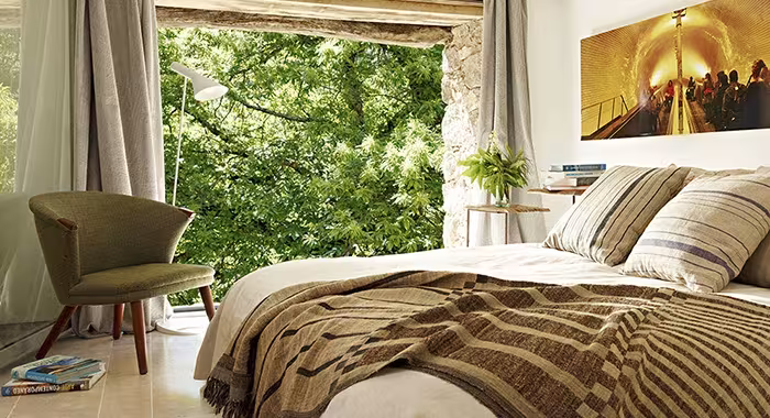 Dormitorio con cama de textiles a rayas, butaca verde y ventanal enmarcando la vegetación del exterior