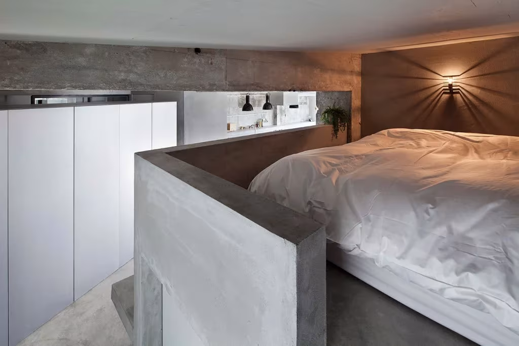 Dormitorio del apartamento de Jiyugaoka con cama blanca tras un murete de microcemento y aplique de pared