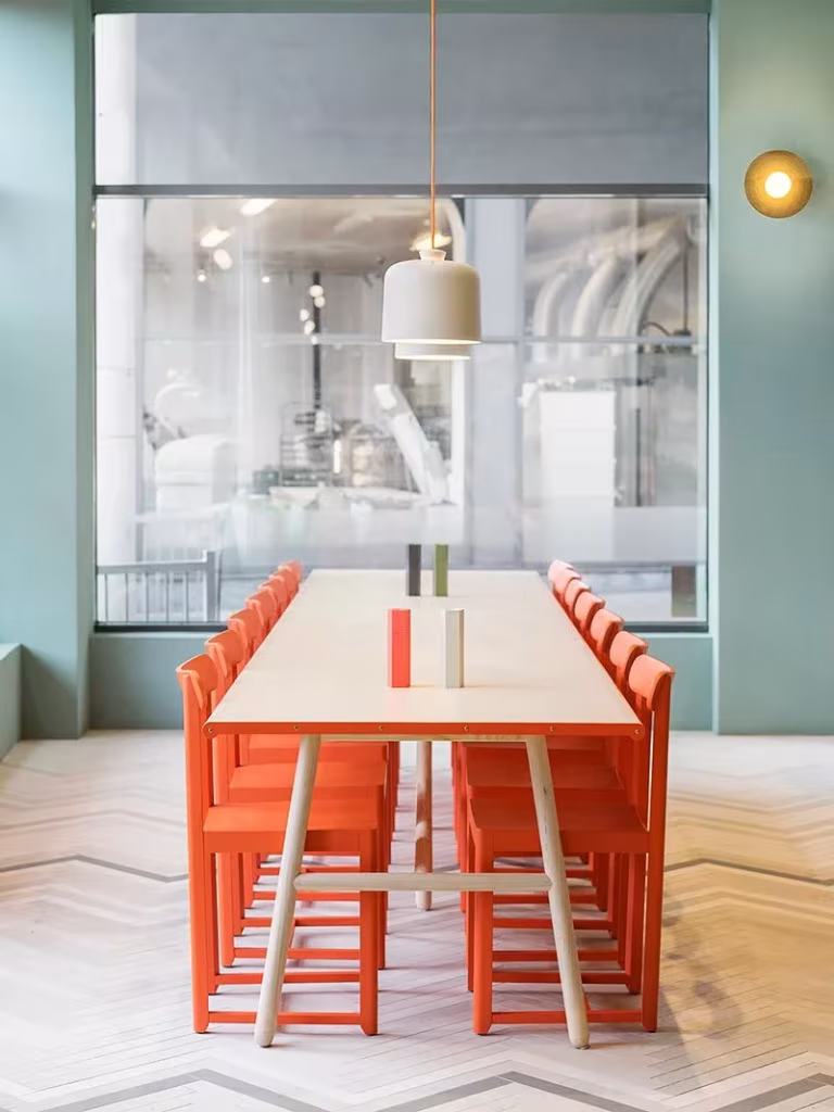 Larga mesa de madera con sillas coral bajo una lámpara colgante frente a la cocina acristalada de Fine Food