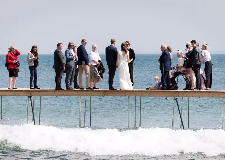 Novios e invitados de boda sobre el puente circular de madera de Aarhus, con olas rompiendo bajo la pasarela