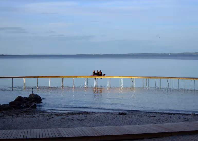 Vista desde la orilla del puente circular de Gjøde & Povlsgaard con personas sentadas sobre el mar al atardecer