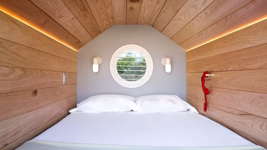 Interior de un habitáculo de siesta revestido de madera, con cama blanca, ojo de buey y teléfono rojo