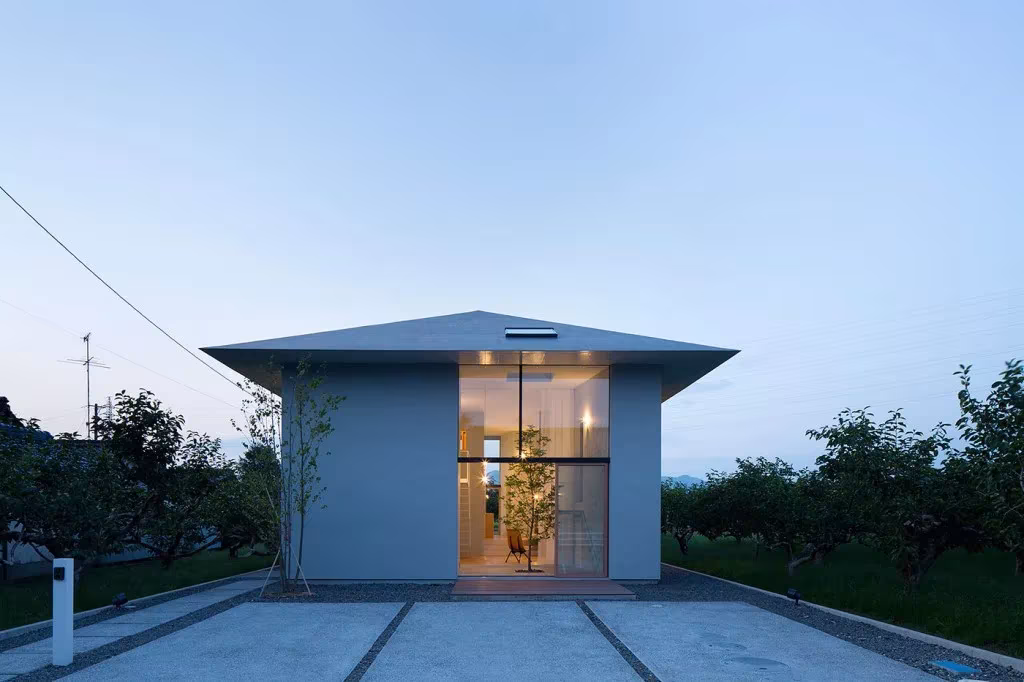 Casa Ohno al anochecer con la entrada acristalada iluminada bajo la cubierta a cuatro aguas entre frutales